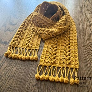 Puede incluir: Una bufanda de ganchillo color amarillo mostaza con un patrón de chevrón repetitivo y un borde de flecos decorado con pequeños pompones redondos. El texto Pattern Digital Download es visible en la esquina inferior derecha de la imagen.