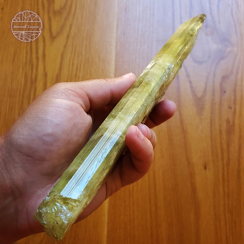 May include: A large, light green, striated crystal point. The crystal is about 10 inches long and has a pointed tip. The crystal is held in a hand, with the background being a wooden surface.