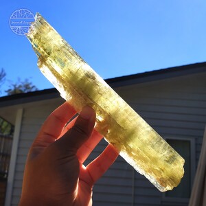 May include: A large, translucent, yellow-green crystal with striations. The crystal is held in a hand against a blue sky background.