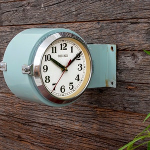 May include: A light blue wall clock with a white face and black numbers. The clock is made by Seiko and has a silver rim. The clock is mounted on a wooden wall.