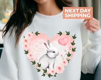 May include: White sweatshirt with a pink heart design featuring a white bunny rabbit and pink roses. The heart is surrounded by green vines.