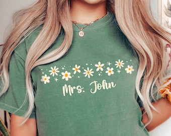 May include: A green t-shirt with a row of white daisies and the text "Mrs. John" in yellow.