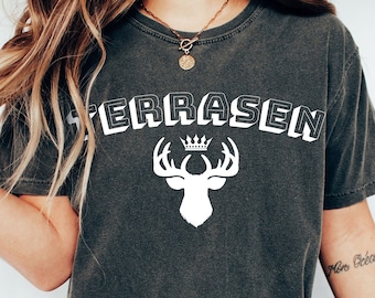 May include: A dark grey t-shirt featuring a white graphic of a deer with antlers and a crown above its head. The text "TERRASEN" is printed in white above the deer. The shirt is being worn by a person wearing blue jeans and white trainers.
