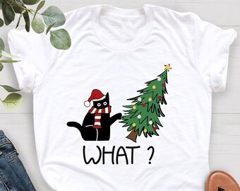 May include: White t-shirt featuring a black cat wearing a Santa hat and scarf, looking at a decorated Christmas tree. The word "WHAT?" is printed below the cat and tree. The shirt is paired with blue jeans and sandals.
