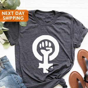 May include: A dark gray t-shirt with a white feminist symbol featuring a raised fist. The symbol is centered on the shirt.