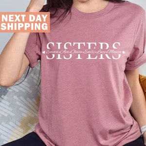 Pode incluir: Uma camiseta malva com a palavra "SISTERS" impressa em branco com os nomes "Emma, Ava, Olivia, Emily, Lucy, Monica" impressos abaixo em um tipo de letra menor.