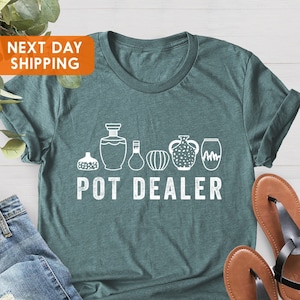 Puede incluir: Una camiseta verde azulada con un gráfico blanco de una fila de plantas en maceta y el texto "POT DEALER".