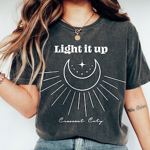 May include: A dark gray t-shirt with a white crescent moon and star graphic. The text "Light it up" and "Crescent City" are printed in white on the shirt.