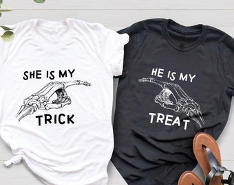 May include: Two t-shirts with a skeleton hand graphic and text. One shirt is white with the text "She is my trick" and the other is black with the text "He is my treat".