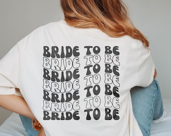 May include: A cream-coloured t-shirt featuring the text "BRIDE TO BE" in a repeating, wavy black font. The design is on the back. The shirt has a loose fit, suitable for casual wear.
