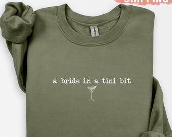 May include: Olive green sweatshirt with the text "a bride in a tini bit" and a martini glass graphic. The long-sleeved top is made of a soft material. A fun, casual top for a bride-to-be.