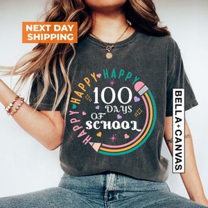 May include: A dark grey t-shirt with a colourful graphic design that says "Happy 100 Days of School" with a rainbow and pencil design. The text "Bella + Canvas" is on the right side of the shirt.