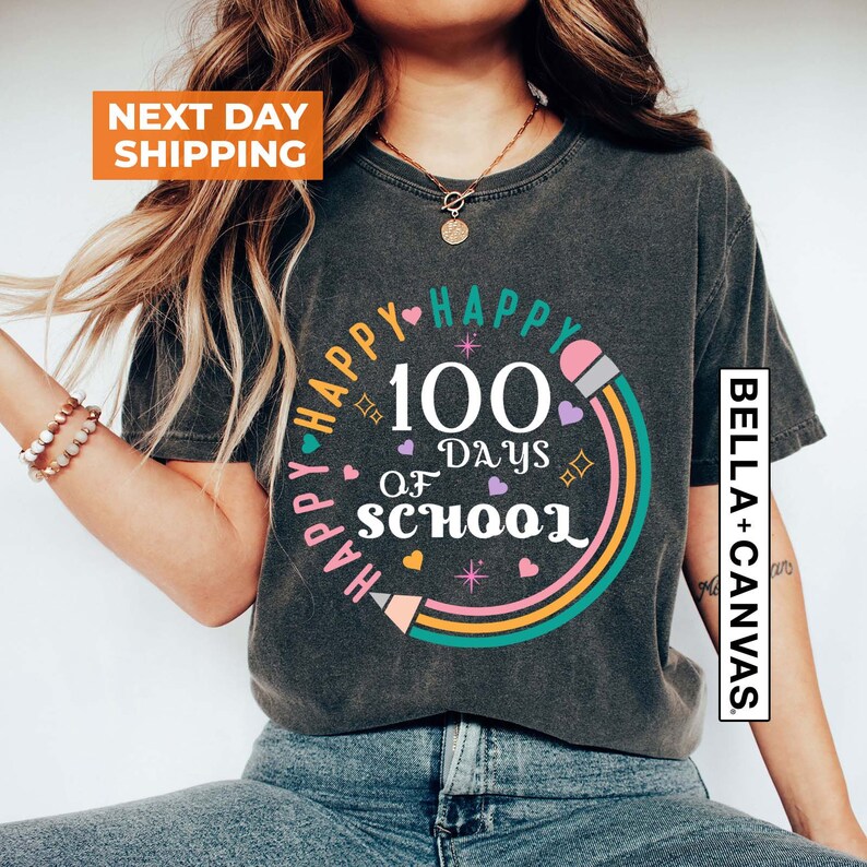 May include: A dark grey t-shirt with a colourful graphic design that says "Happy 100 Days of School" with a rainbow and pencil design. The text "Bella + Canvas" is on the right side of the shirt.
