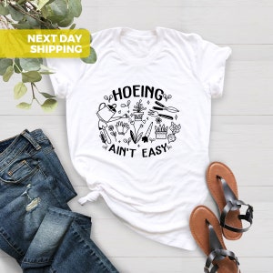 K&ouml;nnte beinhalten: Wei&szlig;es T-Shirt mit einer schwarzen Grafik, auf der "Hoeing ain't easy" steht und verschiedene Gartenwerkzeuge und Pflanzen abgebildet sind.