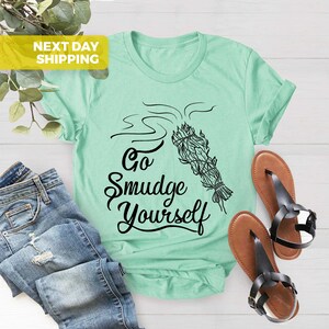 May include: A light green t-shirt with a black graphic of a smudge stick with smoke rising from it. The text "Go Smudge Yourself" is written in a black cursive font.