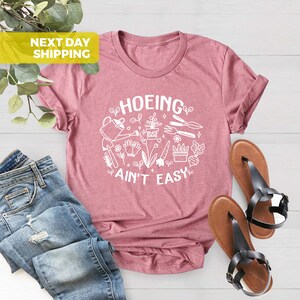 K&ouml;nnte beinhalten: Ein pinkfarbenes T-Shirt mit einer wei&szlig;en Grafik, auf der "HOEING AIN'T EASY" steht, umgeben von Gartenger&auml;ten und Pflanzen.