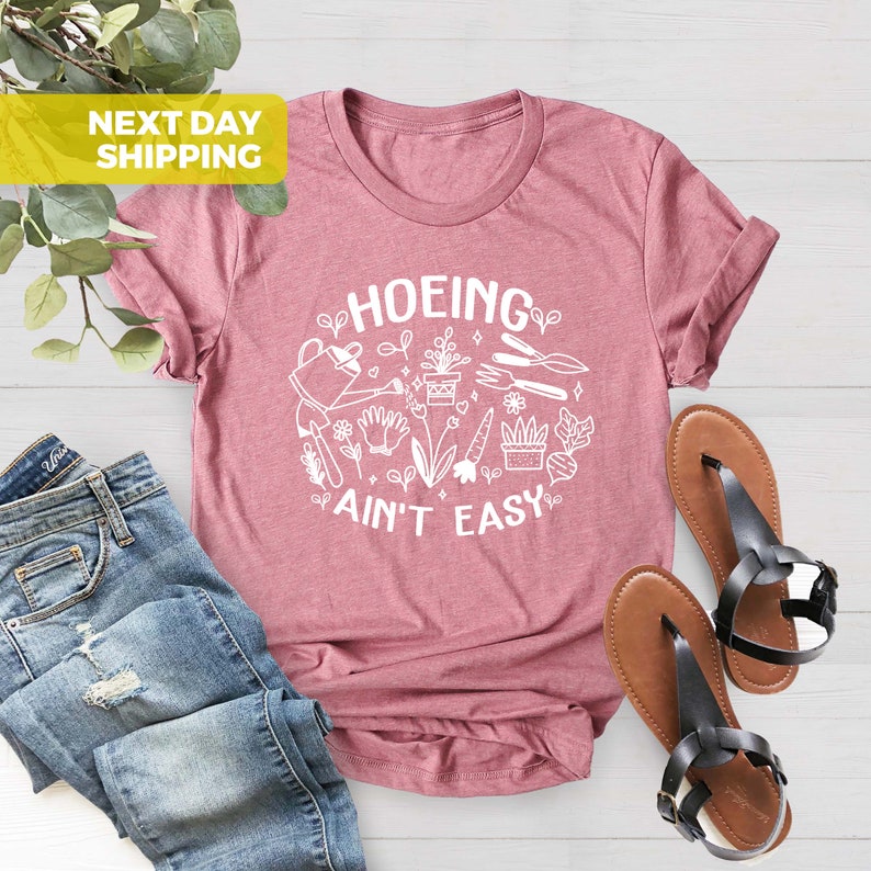 K&ouml;nnte beinhalten: Ein pinkfarbenes T-Shirt mit einer wei&szlig;en Grafik, auf der "HOEING AIN'T EASY" steht, umgeben von Gartenger&auml;ten und Pflanzen.