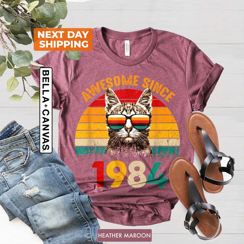 Może przedstawiać: Koszulka w kolorze brązowo-czerwonym z grafiką w stylu vintage przedstawiającą kota w okularach przeciwsłonecznych i tekstem "Awesome Since 1984".