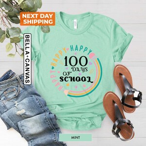 May include: A mint green t-shirt featuring a rainbow design and the text "Happy 100 Days of School" in black. The design includes a pencil, hearts, and stars.