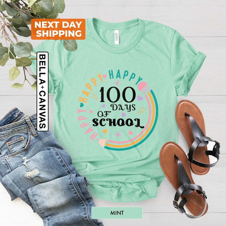 May include: A mint green t-shirt featuring a rainbow design and the text "Happy 100 Days of School" in black. The design includes a pencil, hearts, and stars.