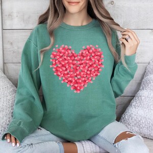 May include: A green sweatshirt with a large heart design made up of smaller pink hearts. The sweatshirt is a relaxed fit and has a crew neck.