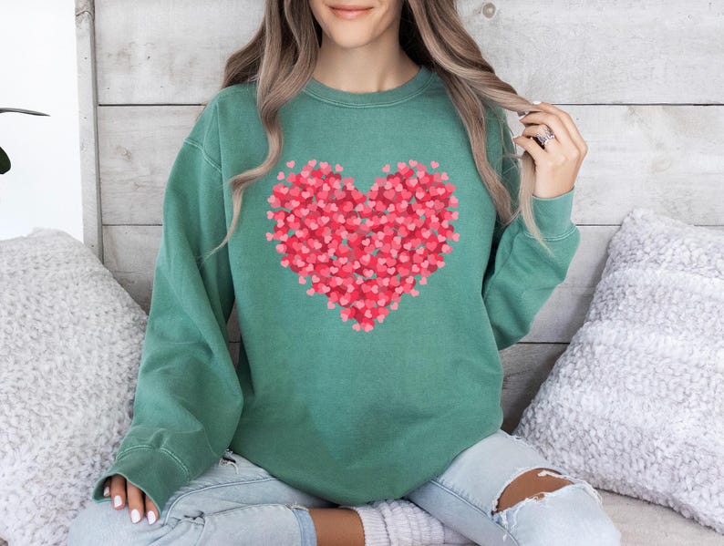 May include: A green sweatshirt with a large heart design made up of smaller pink hearts. The sweatshirt is a relaxed fit and has a crew neck.