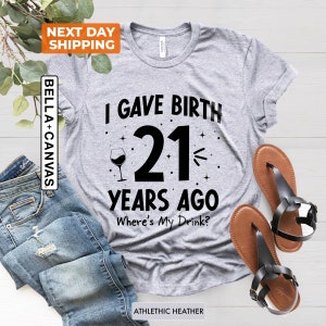 May include: Gray t-shirt with black text that reads "I gave birth 21 years ago. Where's my drink?"  A wine glass with a single glass of wine is to the left of the number 21.  The shirt is on a wood surface.