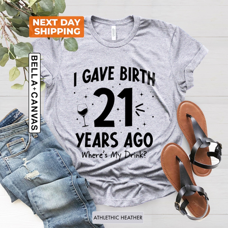 May include: Gray t-shirt with black text that reads "I gave birth 21 years ago. Where's my drink?"  A wine glass with a single glass of wine is to the left of the number 21.  The shirt is on a wood surface.