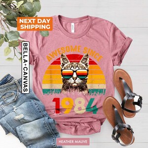 Może przedstawiać: Koszulka w kolorze wrzosowym z vintage'owym nadrukiem kota w okularach przeciwsłonecznych i tekstem "Awesome Since 1984".