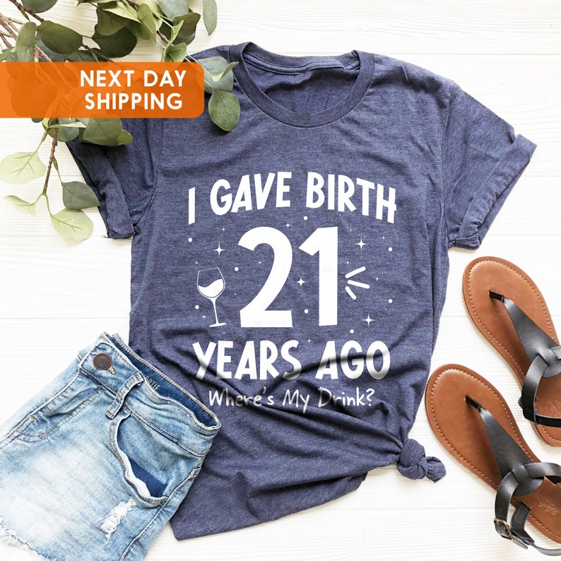 May include: A navy blue t-shirt with white text that reads "I gave birth 21 years ago. Where's my drink?"  A wine glass icon is next to the number 21.