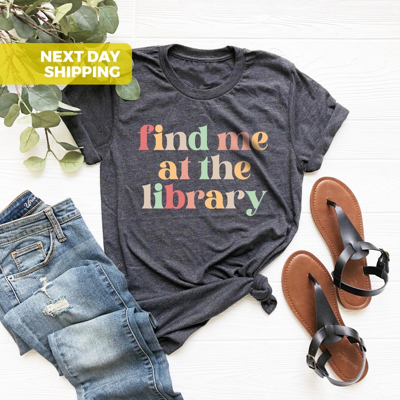 Puede incluir: Una camiseta gris oscuro con el texto "find me at the library" en colores arco&iacute;ris. El texto est&aacute; en un estilo de fuente retro.