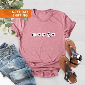 May include: A dusty rose-colored t-shirt with a crew neck and rolled-up sleeves. The shirt features a row of five cartoon sheep in black and white. The text "NEXT DAY SHIPPING" is in an orange box in the upper left corner. Blue jeans and brown sandals are also in the image.