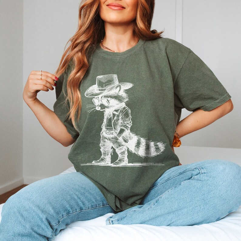 May include: A green t-shirt with a white graphic of a raccoon wearing a cowboy hat and sunglasses. The raccoon is standing with its hands on its hips.