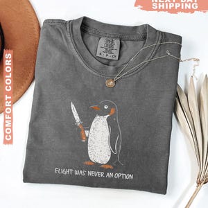 May include: A charcoal grey t-shirt featuring a graphic of a penguin holding a knife with the text "FLIGHT WAS NEVER AN OPTION" printed below. The shirt has a crew neckline and a Comfort Colors brand label on the inside of the collar area.