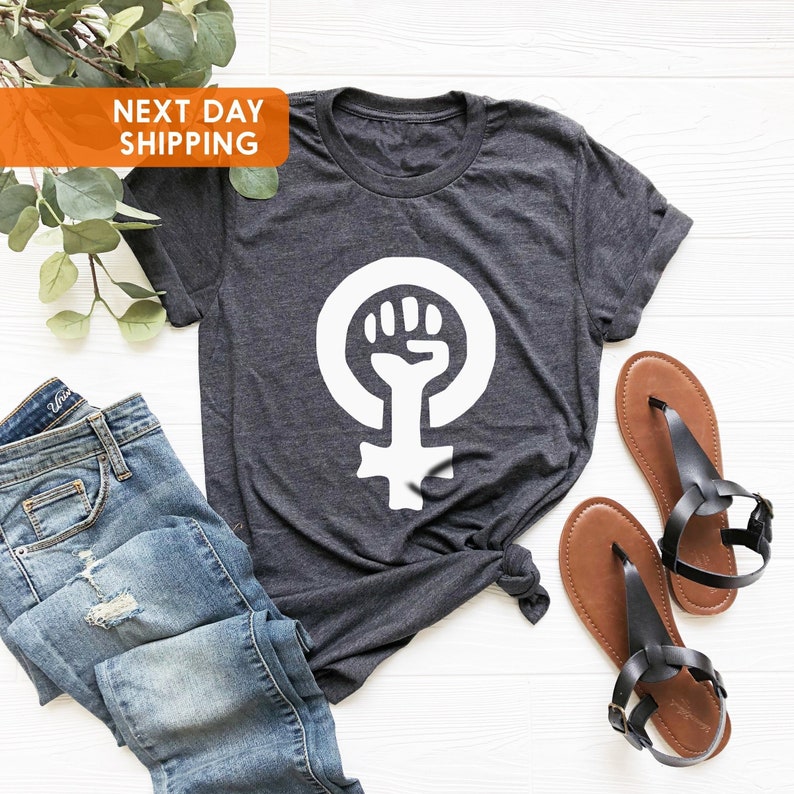 May include: A dark gray t-shirt with a white feminist symbol featuring a raised fist. The symbol is centered on the shirt.