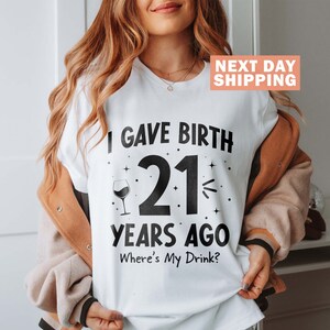 May include: White t-shirt with black text that reads "I gave birth 21 years ago. Where's my drink?" and a black silhouette of a wine glass.