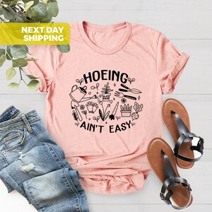 K&ouml;nnte beinhalten: Ein pinkfarbenes T-Shirt mit einem schwarzen Grafikdesign, auf dem "HOEING AIN'T EASY" steht, umgeben von Gartenger&auml;ten und Pflanzen.
