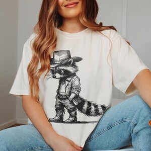 May include: White t-shirt with a black and white illustration of a raccoon wearing a cowboy hat and sunglasses. The raccoon is holding a gun and has a bandana around its neck.