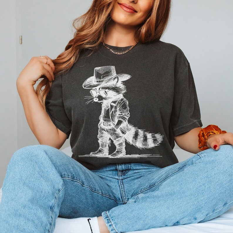 May include: A black t-shirt with a white graphic of a raccoon wearing a cowboy hat and holding a gun. The raccoon is wearing a jacket and boots.