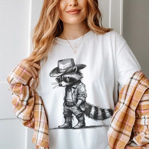 May include: A white t-shirt with a black and white illustration of a raccoon wearing a cowboy hat and sunglasses. The raccoon is standing with its hands on its hips and has a long tail.