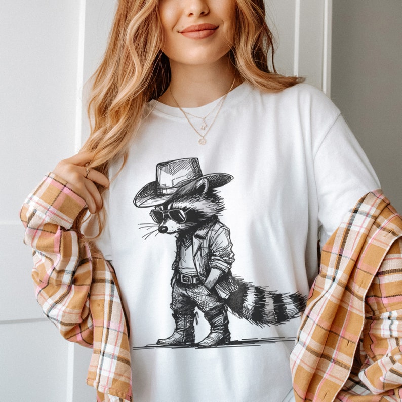 May include: A white t-shirt with a black and white illustration of a raccoon wearing a cowboy hat and sunglasses. The raccoon is standing with its hands on its hips and has a long tail.