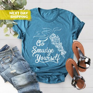 May include: Teal blue t-shirt with white text that says "Go Smudge Yourself" and a white illustration of a smudge stick with smoke coming from it.