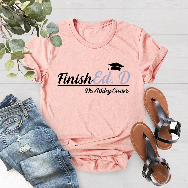 Puede incluir: Una camiseta rosa con el texto "Finish Ed. D" en negro con un icono de gorro de graduaci&oacute;n sobre el texto. El texto "Dr. Ashley Carter" est&aacute; debajo del texto principal.