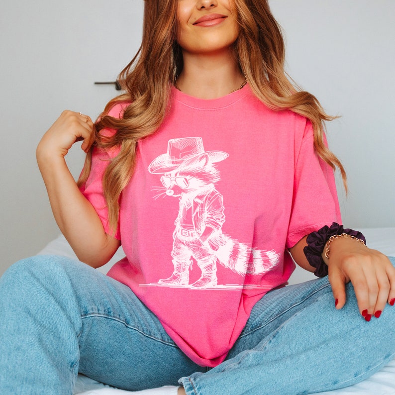 May include: A pink t-shirt with a white graphic of a raccoon wearing a cowboy hat and sunglasses. The raccoon is standing with its arms crossed and has a striped tail.