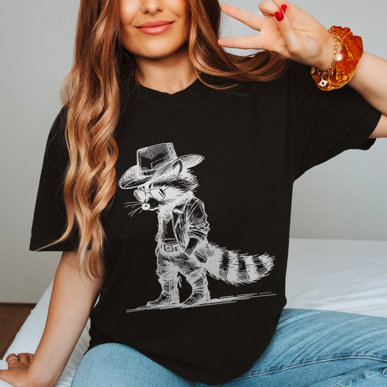May include: A black t-shirt with a white graphic of a raccoon wearing a cowboy hat and sunglasses. The raccoon is wearing a leather jacket and jeans.