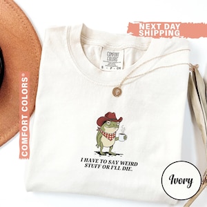 May include: Ivory-colored t-shirt featuring a cartoon frog wearing a cowboy hat and holding a coffee cup. The frog has a red and white checkered bandana. The text "I HAVE TO SAY WEIRD STUFF OR I'LL DIE" is printed below the frog.