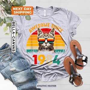 Może przedstawiać: Szara koszulka z grafiką w stylu vintage przedstawiającą kota w okularach przeciwsłonecznych i tekstem "Awesome Since 1984".