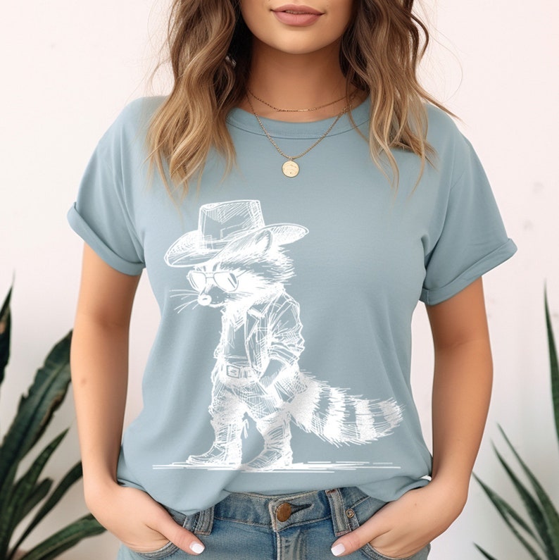 May include: A light blue t-shirt with a white graphic of a raccoon wearing a cowboy hat and sunglasses. The raccoon is wearing a jacket and boots.