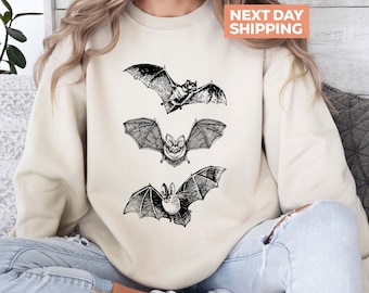 May include: Cream-colored sweatshirt featuring three detailed black bat illustrations. The bats are depicted in various poses, with outstretched wings. The sweatshirt has a crew neck and long sleeves. The text "NEXT DAY SHIPPING" is in a peach-colored box.