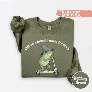 May include: Military green sweatshirt with the text "Off to commit mom foolery." The graphic features a cartoon frog wearing a witch hat and riding a scooter. The sweatshirt is a crew neck style.
